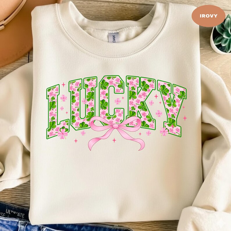 May include: Cream-colored sweatshirt with the word "LUCKY" in green, floral-filled letters. The letters are outlined in green and filled with pink flowers and green shamrocks. A pink bow is centered below the word, with pink star accents.