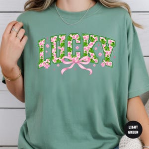 May include: Light green t-shirt with the word "LUCKY" in green block letters filled with a pink floral and shamrock pattern. A pink bow is centered below the word, with pink flowers scattered around.