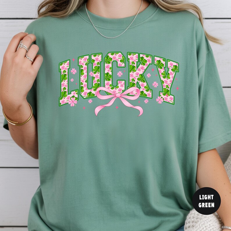 May include: Light green t-shirt with the word "LUCKY" in green block letters filled with a pink floral and shamrock pattern. A pink bow is centered below the word, with pink flowers scattered around.
