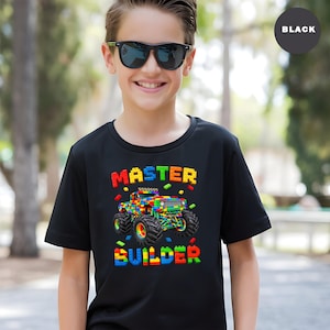 May include: Black t-shirt with a colorful graphic of a monster truck made of toy blocks. The words "MASTER BUILDER" are displayed above and below the truck in block letters. The shirt is worn by a person wearing sunglasses.