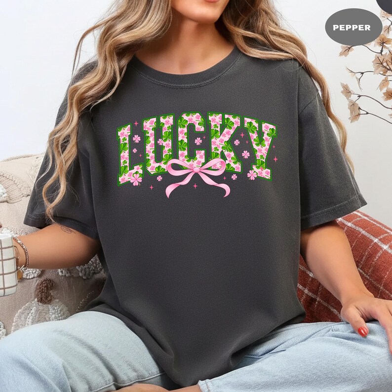 May include: Dark gray t-shirt with the word "LUCKY" in a green floral design with pink accents and a pink bow. The shirt is a casual style, suitable for everyday wear.
