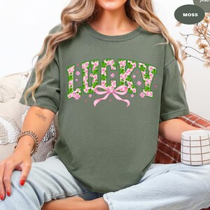 May include: Moss green t-shirt with the word "LUCKY" in a floral design. The letters are filled with green shamrocks and pink flowers, with a pink bow below. A St. Patrick's Day themed tee.