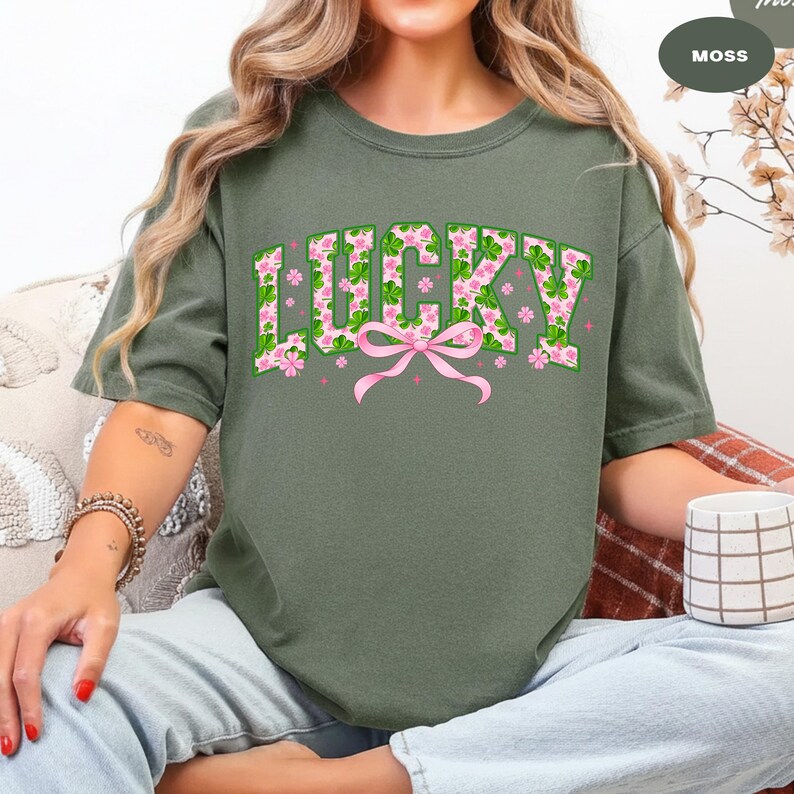 May include: Moss green t-shirt with the word "LUCKY" in a floral design. The letters are filled with green shamrocks and pink flowers, with a pink bow below. A St. Patrick's Day themed tee.