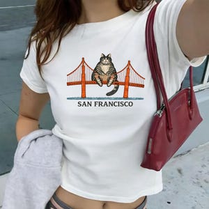 May include: White t-shirt featuring a cartoon cat sitting on the Golden Gate Bridge in San Francisco. The bridge is depicted in orange with a blue base, and the text "SAN FRANCISCO" is below the image.