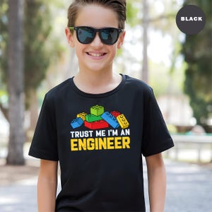 May include: Black t-shirt with a colorful graphic of toy building blocks and the text "TRUST ME I'M AN ENGINEER" in yellow and white. The shirt is worn by a person wearing sunglasses.