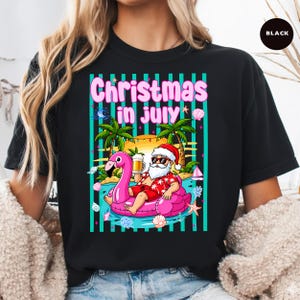 May include: A black t-shirt featuring a graphic print with the text Christmas in July. The design shows Santa Claus wearing sunglasses and a red floral shirt, sitting on a pink flamingo pool float while holding a beer mug on a beach.