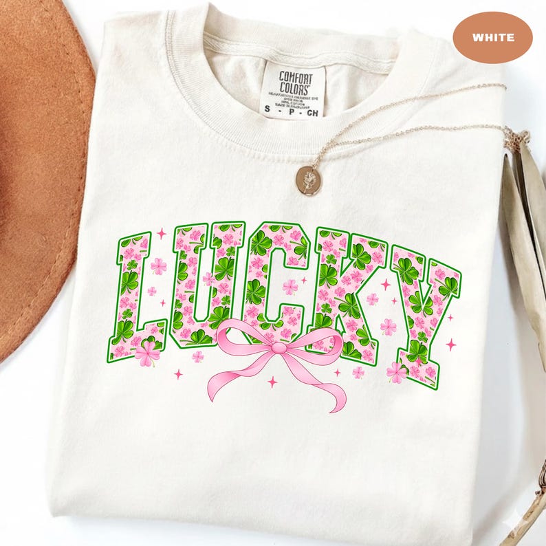 May include: White t-shirt with the word "LUCKY" in green outlined letters filled with pink flowers and green shamrocks. A pink bow is below the word. The shirt has a crew neck and short sleeves. A gold necklace is draped across the shirt.