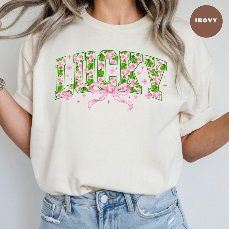 May include: Cream-colored sweatshirt with the word "LUCKY" in green block letters filled with pink flowers and shamrocks. A pink bow is centered below the word. The sweatshirt is a casual style.