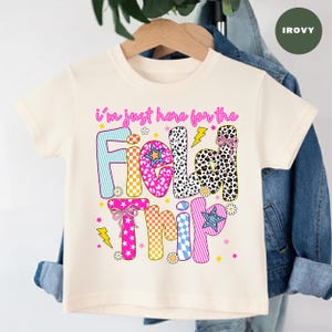 May include: Cream-colored short-sleeved t-shirt with the phrase "I'm just here for the Field Trip" in colorful, patterned letters. The design includes stars, lightning bolts, and floral accents, creating a playful and vibrant look.