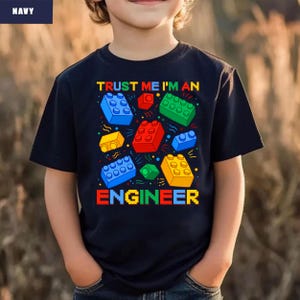 May include: Navy blue t-shirt with the text "TRUST ME I'M AN ENGINEER" in colorful pixelated letters. The shirt features a design of colorful toy building blocks in red, blue, green, and yellow.