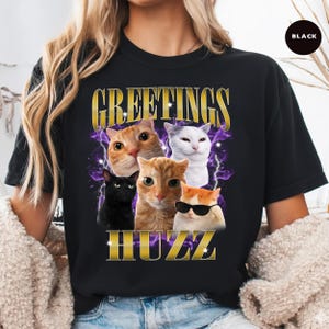 May include: Black t-shirt featuring a graphic of five cats with the text "GREETINGS HUZZ" in gold lettering. The cats are in various colors, including orange, white, and black, with one wearing sunglasses. Purple lightning bolts are in the background.