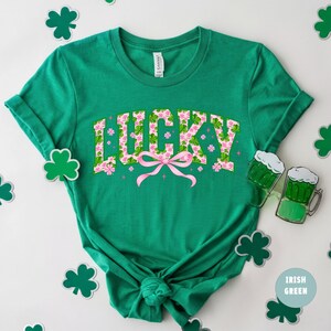 May include: A green t-shirt with the word "LUCKY" in a floral design with a pink bow. The shirt is surrounded by shamrock cutouts and two green beer mugs. The text "Irish Green" is also visible.