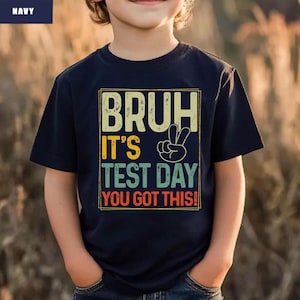 May include: Navy blue t-shirt with the text "BRUH IT'S TEST DAY YOU GOT THIS!" in a retro style. The design includes a peace sign. The shirt is a short-sleeved crew neck.