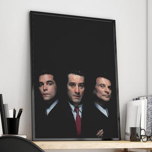 May include: Framed poster featuring three men in suits against a black background. The central figure wears a red tie. The poster is displayed on a white wall, with office supplies and books visible in the background.