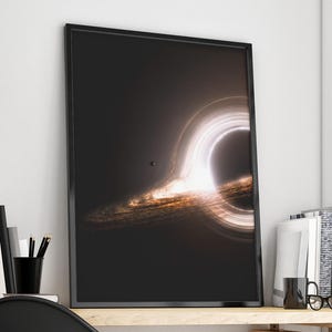 Op de afbeelding: Ingelijste kunstdruk met een zwart gat met een heldere, wervelende accretieschijf. Het kunstwerk heeft een donkere achtergrond met een gloeiend, cirkelvormig licht en een kleine, donkere bol. De print zit in een zwart frame.