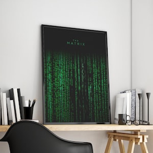 May include: A framed poster displaying the green digital rain from "The Matrix". The film's title, "THE MATRIX", is in green at the top against a black backdrop. The artwork is displayed on a wooden desk, with books and a black chair.
