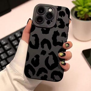 May include: A dark gray phone case with a black leopard print design. The case has a camera lens protector and is held in a hand. The background includes a keyboard and a small potted plant.