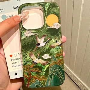 May include: Green phone case with a landscape design featuring cranes, clouds, a yellow sun, and mountains. The case has a glossy finish and a cutout for the camera. The design is inspired by traditional Asian art.