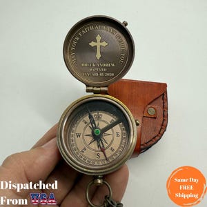 May include: A vintage-style brass compass with a leather case. The compass lid is engraved with a cross and the text "MAY YOUR FAITH ALWAYS GUIDE YOU". The compass face displays cardinal directions and degree markings. The text "BRECK ANDREW BAPTIZED JANUARY 18, 2026" is also engraved.