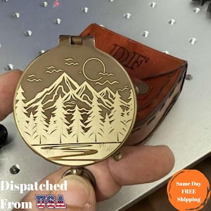 May include: A bronze-colored compass with a mountain and forest design, held near a brown leather case. The compass features a detailed landscape engraving with trees, mountains, a sun, and water. The text "Dispatched From USA" is visible at the bottom.
