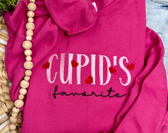 May include: A bright pink sweatshirt with the text "CUPID'S favorite" in white and black. Red heart accents are above the word "favorite". The sweatshirt is folded on top of a pair of light blue jeans.