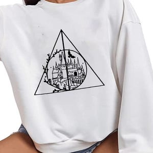 May include: White sweatshirt with a black graphic of a castle inside a triangle. The long-sleeved top has a crew neck and a relaxed fit. The graphic includes a castle, a bird, and floral elements. The sweatshirt is made of a soft material.