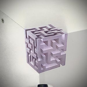 May include: A three-dimensional, lavender-colored cube with a complex maze design. The cube appears to be floating in the corner of a white room, creating an optical illusion. The artwork is a geometric puzzle.