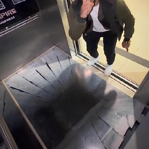 May include: An optical illusion of a deep hole in the floor. The floor is made of gray tiles with cracks. A person is standing near the hole, pointing a finger to their mouth.