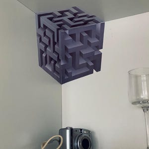 Puede incluir: Un diseño de laberinto cúbico gris tridimensional está fijado a una esquina blanca. Una cámara digital plateada y un vaso transparente están en un estante blanco debajo. El diseño del laberinto parece flotar.