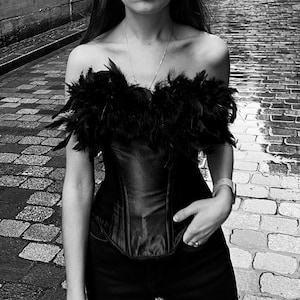 May include: A black corset top with a strapless design, adorned with black feathers along the neckline. The top is paired with black pants. The image is in black and white, showcasing the texture of the clothing.