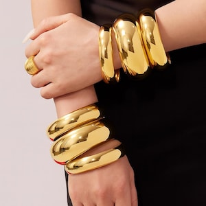 May include: Gold-colored jewelry set featuring multiple wide, shiny bangles and rings. The bangles are worn on both wrists, and the rings are on the fingers. The jewelry has a polished, reflective surface, creating a bold and glamorous look.