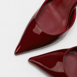 May include: A pair of burgundy-colored, pointed-toe pumps. The shoes have a glossy patent leather finish with a red trim around the opening. The image is a close-up shot of the shoes against a white background.