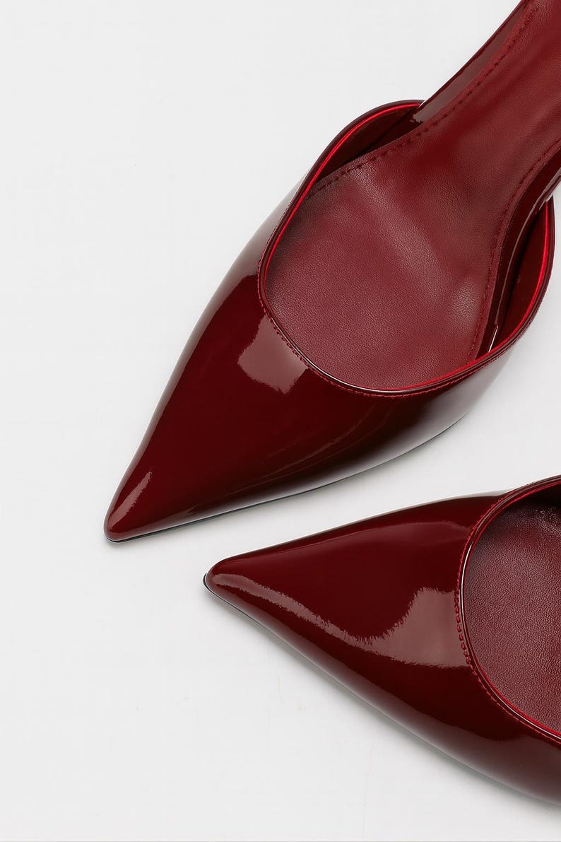 May include: A pair of burgundy-colored, pointed-toe pumps. The shoes have a glossy patent leather finish with a red trim around the opening. The image is a close-up shot of the shoes against a white background.