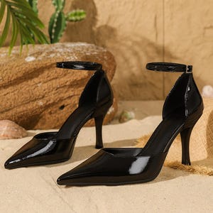 May include: Pair of black patent leather high-heeled shoes with pointed toes and ankle straps. The stiletto heels are slender and the shoes have a glossy finish. The shoes are displayed on a sandy surface.