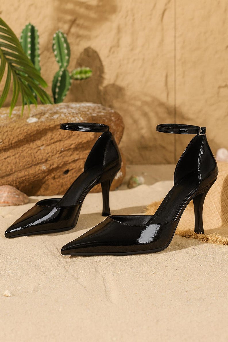 May include: Pair of black patent leather high-heeled shoes with pointed toes and ankle straps. The stiletto heels are slender and the shoes have a glossy finish. The shoes are displayed on a sandy surface.