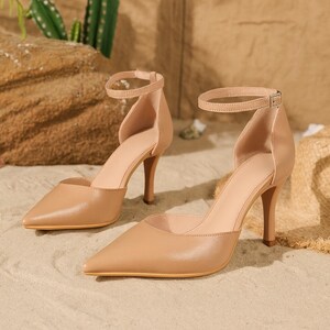 May include: A pair of beige high-heeled shoes with pointed toes and ankle straps. The heels are approximately 3 inches tall. The shoes are made of a smooth material and are set against a sandy background.
