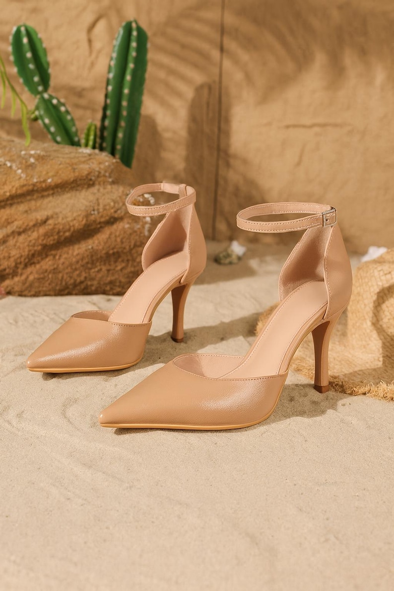 May include: A pair of beige high-heeled shoes with pointed toes and ankle straps. The heels are approximately 3 inches tall. The shoes are made of a smooth material and are set against a sandy background.