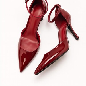 May include: A pair of red patent leather high heels with pointed toes and ankle straps. The shoes have a stiletto heel and a glossy finish, set against a white background. The heels are approximately 3 inches high.
