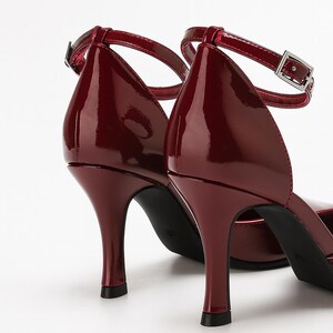 May include: A pair of burgundy patent leather high heels with ankle straps. The shoes have a pointed toe and a thin, curved heel. The straps have a silver buckle closure. The shoes are set against a white background.