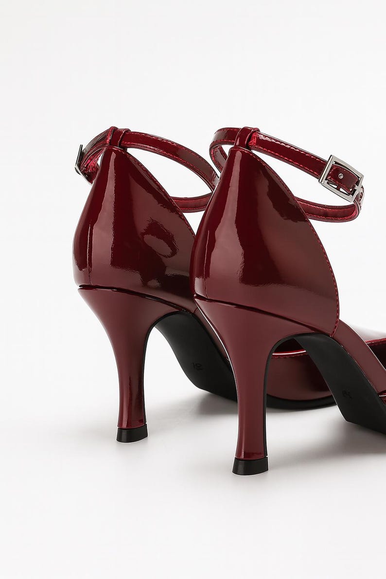 May include: A pair of burgundy patent leather high heels with ankle straps. The shoes have a pointed toe and a thin, curved heel. The straps have a silver buckle closure. The shoes are set against a white background.
