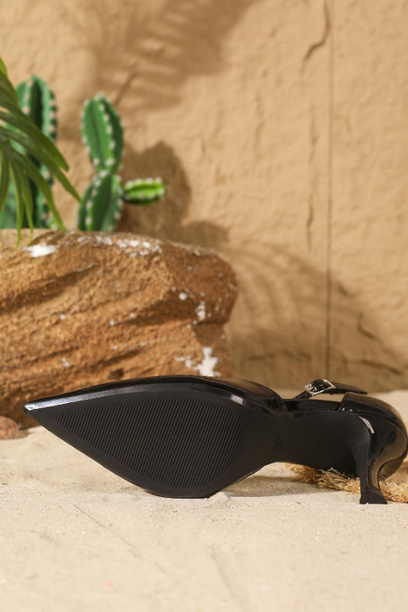 May include: Black patent leather pointed-toe pump with a delicate ankle strap and a stiletto heel. The shoe features a textured sole and a sleek, modern design. The heel height is approximately 3 inches.