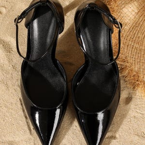 May include: Pair of black patent leather pumps with pointed toes and ankle straps. The shoes have a glossy finish and are set against a sandy background, suggesting a beach or outdoor setting. The heels are not visible.
