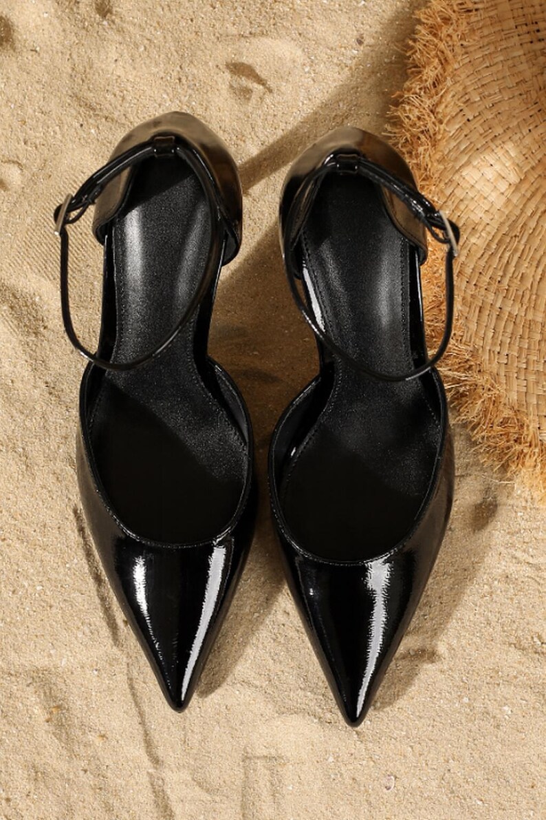 May include: Pair of black patent leather pumps with pointed toes and ankle straps. The shoes have a glossy finish and are set against a sandy background, suggesting a beach or outdoor setting. The heels are not visible.