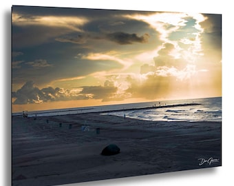 May include: A framed print of a beach scene at sunset. The sky is filled with dramatic clouds, with golden sunlight breaking through. The beach is sandy, and the ocean is visible in the distance. A few people are seen on the pier.