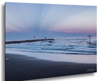May include: A scenic photograph of a beach at dusk. The sky transitions from light blue to pink and purple. A stone pier extends into the ocean, with a few figures visible. Waves gently roll onto the shore. A sign stands in the water.