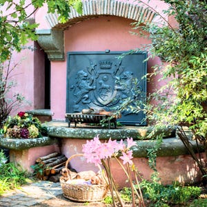 Puede incluir: Una pantalla decorativa de chimenea de metal oscuro con un diseño detallado de escudo de armas, flanqueado por leones y otros elementos heráldicos. La pantalla está colocada contra una pared de estuco rosa, con un pequeño banco decorativo en frente. Una cesta con cachorros y flores rosas está en primer plano.