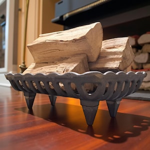 Op de afbeelding: Een zwarte, gietijzeren haardhoutmand met een geschulpte rand en vier puntige poten. De houder bevat verschillende stukken lichtbruin brandhout. De houder staat op een donkerhouten oppervlak.