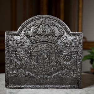 Op de afbeelding: Een donkergrijze gietijzeren haardplaat met een gewelfde bovenkant. Het ontwerp toont een gedetailleerd reliëfontwerp van een kroon, adelaars en een wapenschild. Het algehele ontwerp is sierlijk en decoratief, geschikt voor een traditioneel of antiek huis.