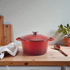 Può includere: Una casseruola in ghisa smaltata rossa con coperchio e manici laterali. È appoggiata su una superficie di legno con un cucchiaio di legno, un panno bianco piegato e una piccola pianta in vaso. Un elemento essenziale in cucina.