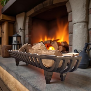 Op de afbeelding: Een donkergrijze gietijzeren haardhoutdrager gevuld met brandhout. De houder heeft een rechthoekige vorm met afgeronde randen en staat op drie korte poten. Een open haard met een brandend vuur is op de achtergrond.